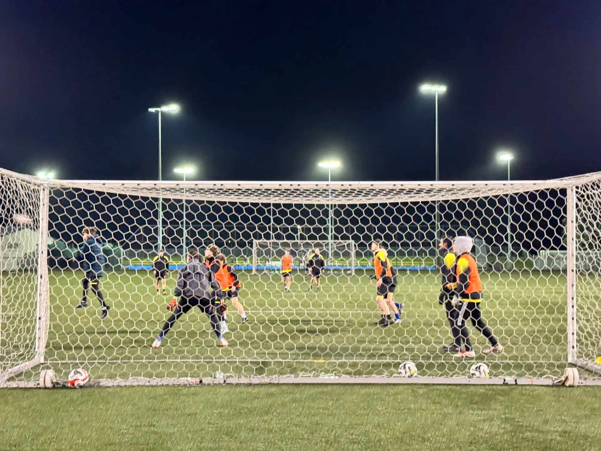 Club footballers training under floodlights at night, seen through the goal net
